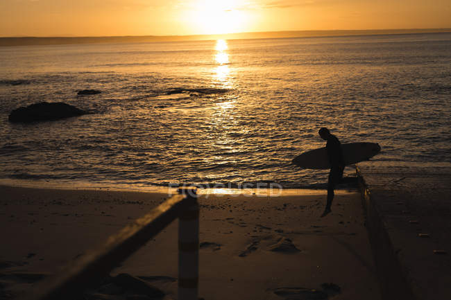 Surfista con tavola da surf in spiaggia durante il tramonto — Foto stock