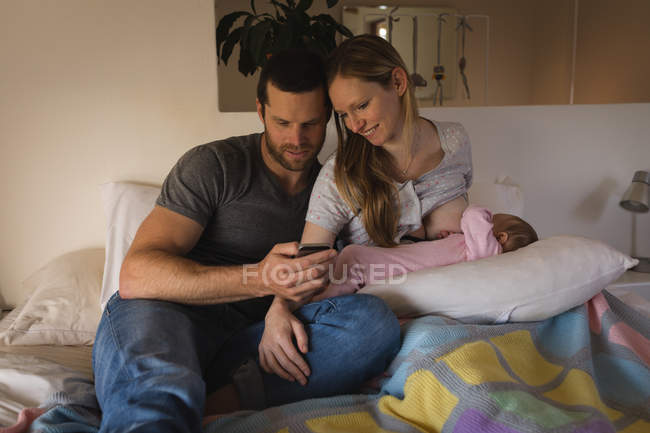 Eltern nutzen Mobiltelefon, während die stillende Mutter ihr Baby zu Hause im Bett hält — Stockfoto