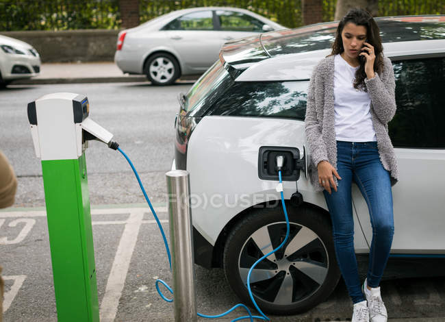 Mujer hablando por teléfono móvil mientras carga coche eléctrico en la estación de carga - foto de stock