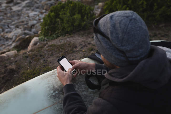 Vue grand angle de l'homme avec planche de surf en utilisant mobile sur la plage — Photo de stock