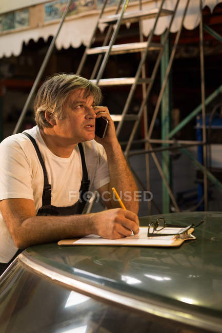Мужчина-механик разговаривает по мобильному телефону во время записи в буфер обмена в гараже — стоковое фото