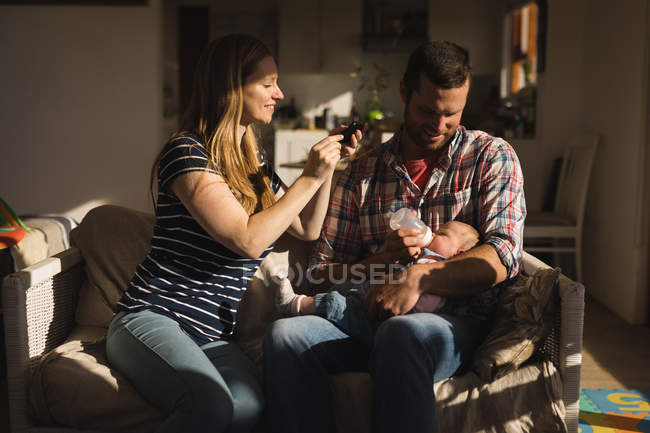 Madre tomando fotos desde el móvil mientras su padre alimenta a su bebé en el sofá en casa - foto de stock
