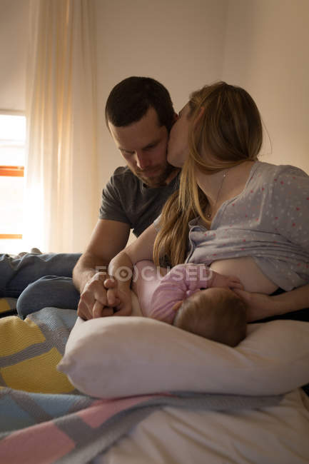 Родители целуются друг с другом, пока мать кормит грудью ребенка в постели дома — стоковое фото