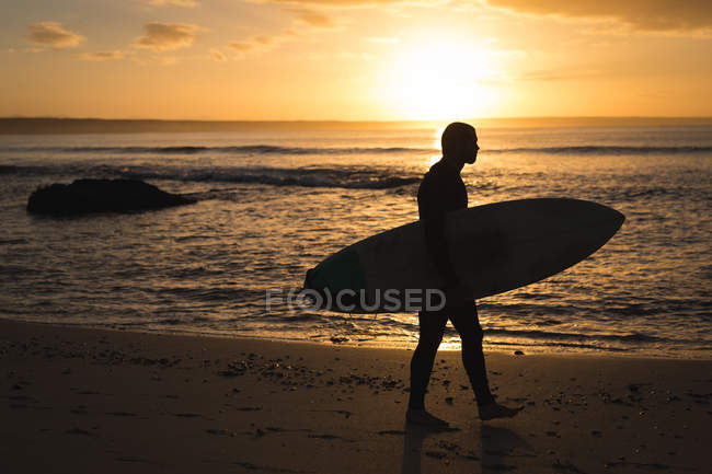 Surfista con tabla de surf caminando por la playa al atardecer - foto de stock