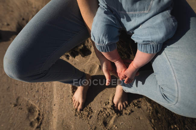Sección baja de bebé sentado en madres regazo en la playa - foto de stock