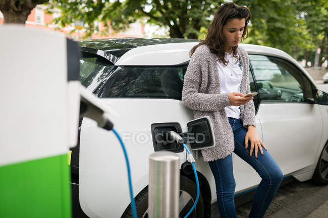 Женщина с мобильного телефона во время зарядки электромобиля на зарядной станции — стоковое фото