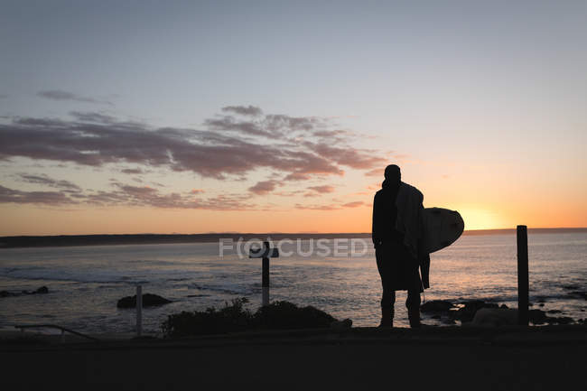 Вид ззаду людини з дошкою для серфінгу, що стоїть на пляжі під час заходу сонця — стокове фото