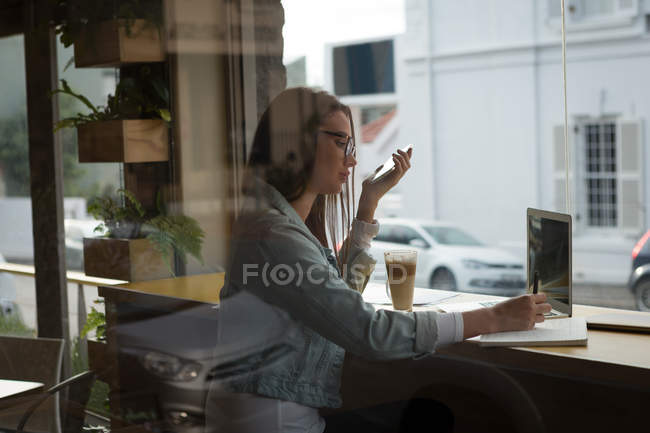 Donna che scrive sul diario mentre parla sul cellulare nel caffè — Foto stock
