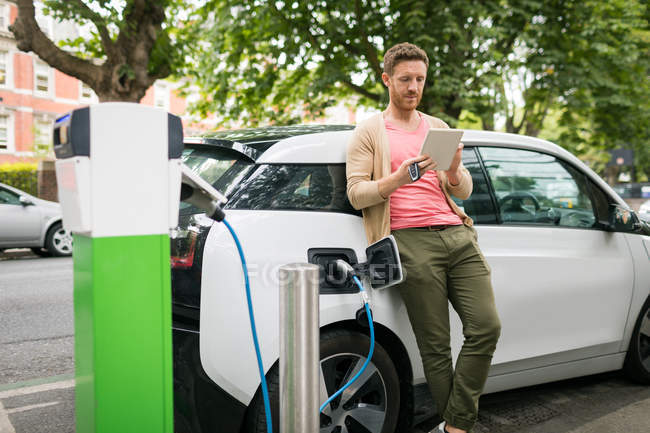 Man using digital tablet while charging electric car at charging station — Stock Photo