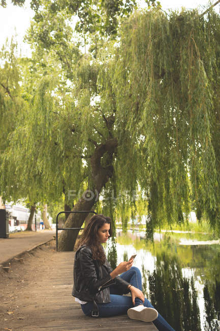 Женщина пользуется мобильным телефоном, сидя у берега озера — стоковое фото