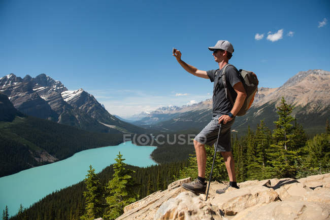 Hombre haciendo clic en imágenes con el teléfono móvil, mientras que de pie en una roca en el campo - foto de stock