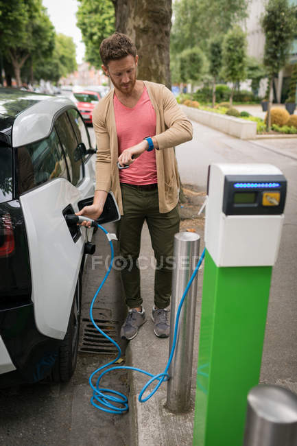 Man checking time on smart watch while charging electric car at charging station — Stock Photo