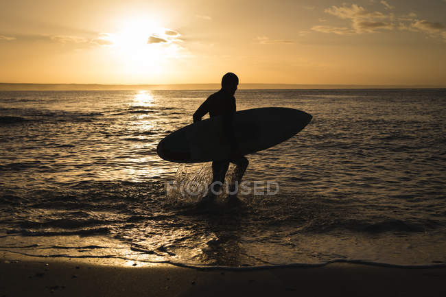 Surfista con tabla de surf caminando en la playa al atardecer - foto de stock
