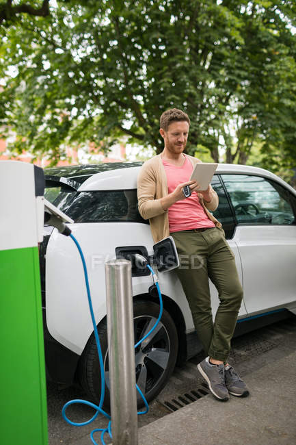 Man using digital tablet while charging electric car at charging station — Stock Photo