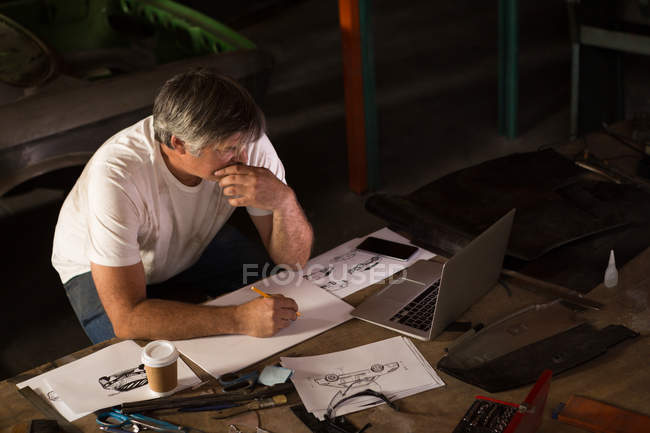 Male mechanic drawing a car model while using laptop in garage — Stock Photo