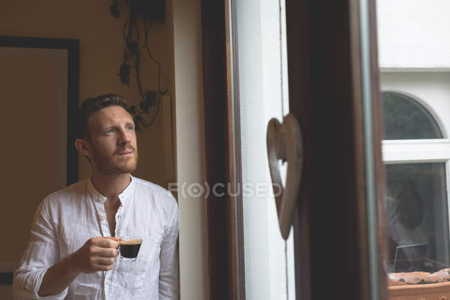 Задумчивый человек пьет черный кофе, стоя у окна дома — стоковое фото