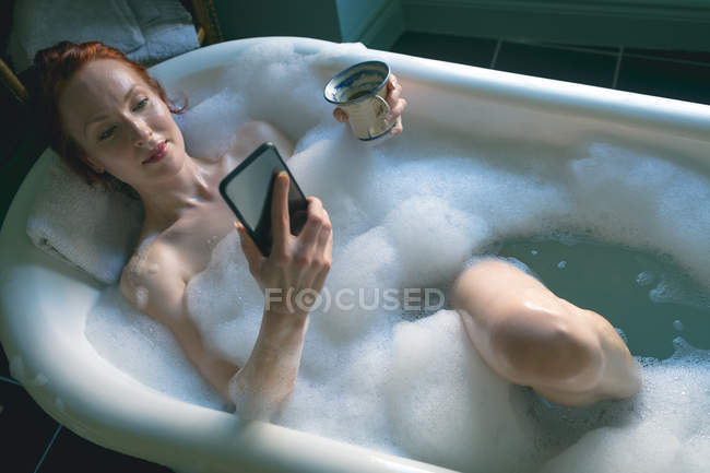 Жінка використовує мобільний телефон з чашкою кави у ванній кімнаті — стокове фото