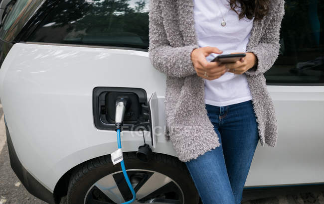 Средняя секция женщины, использующей мобильный телефон при зарядке электромобиля на зарядной станции — стоковое фото