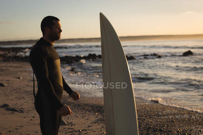 Surfista com prancha de surf em pé na praia durante o pôr do sol — Fotografia de Stock