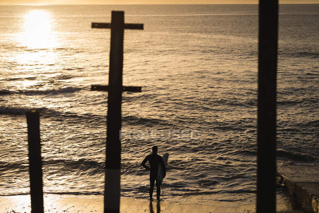 Surfista com prancha de surf em pé na praia durante o pôr do sol — Fotografia de Stock