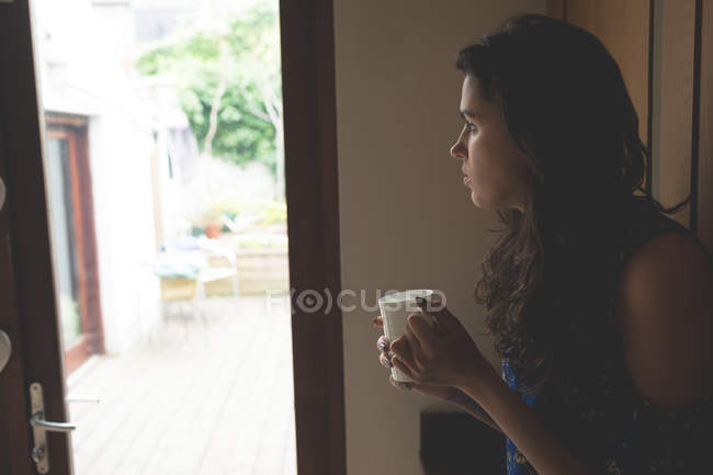 Задумчивая женщина пьет кофе, стоя у окна дома — стоковое фото