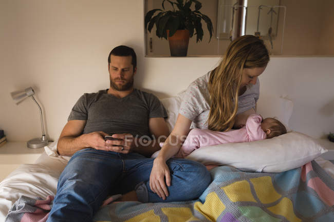 Отец с помощью мобильного телефона, пока мать кормит грудью ребенка на кровати дома — стоковое фото