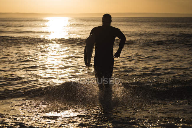 Surfista com prancha correndo para o mar durante o pôr do sol — Fotografia de Stock