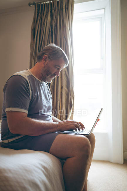 Пожилой мужчина использует ноутбук в спальне дома — стоковое фото