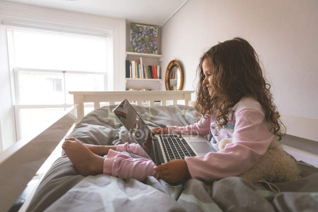 Chica usando el ordenador portátil en la cama en el dormitorio en casa - foto de stock