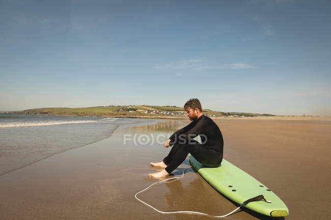 Vista lateral do surfista sentado na prancha de surf na praia — Fotografia de Stock