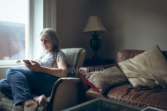 Mujer mayor usando teléfono móvil en la sala de estar en casa - foto de stock