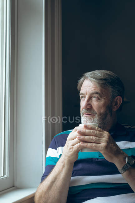 Продуманий старший чоловік дивиться крізь вікно, маючи каву вдома — стокове фото