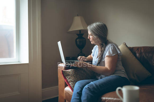 Mulher sênior usando laptop na sala de estar em casa — Fotografia de Stock