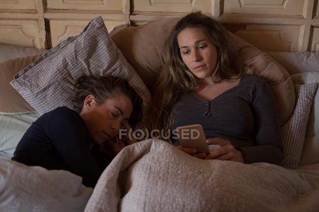 Pareja lesbiana relajándose y utilizando el teléfono móvil en la cama en casa - foto de stock