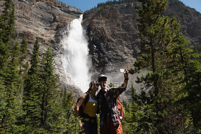 Casal tirando selfie com telefone celular perto de cachoeira — Fotografia de Stock