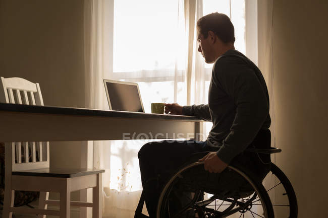 Людина з обмеженими можливостями має каву під час використання ноутбука на обідньому столі вдома — стокове фото