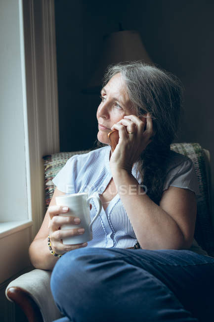 Senior woman talking on mobile phone while having coffee at home — Stock Photo