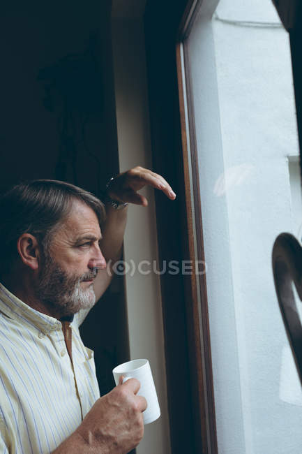 Продуманий старший чоловік з чашкою кави дивиться через вікно вдома — стокове фото