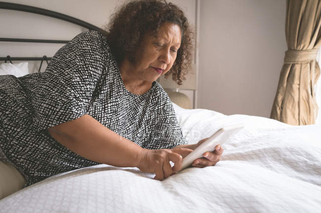 Пожилая женщина с цифровым планшетом на кровати в спальне дома — стоковое фото