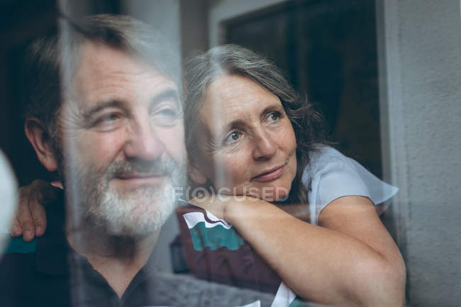 Thoughtful senior couple looking through window at home — Stock Photo