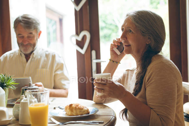 Старшая женщина разговаривает по мобильному телефону за чашечкой кофе дома — стоковое фото