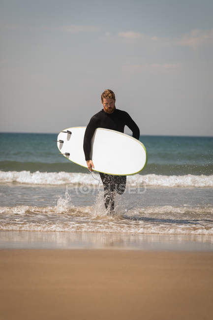 Surfista com prancha de surf correndo na praia no dia ensolarado — Fotografia de Stock