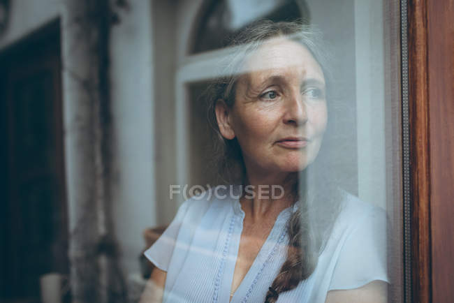 Femme âgée réfléchie regardant par la fenêtre à la maison — Photo de stock