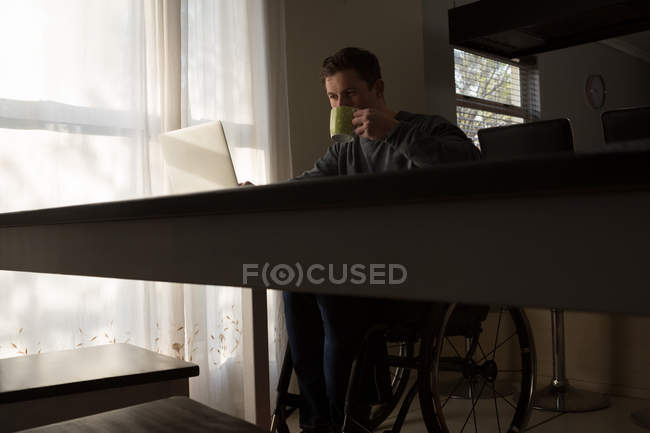 Hombre discapacitado tomando café mientras usa el portátil en la mesa de comedor en casa - foto de stock