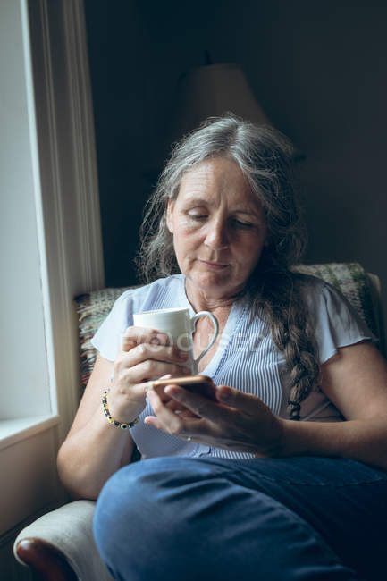 Старша жінка використовує мобільний телефон, маючи каву вдома — стокове фото