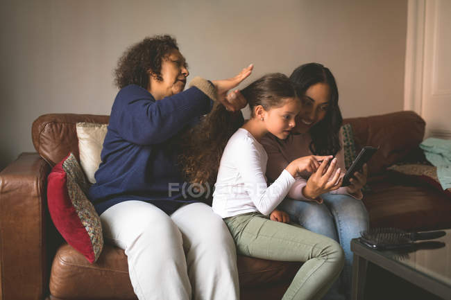 Бабуся робить зачіску онуки, використовуючи мобільний телефон з матір'ю вдома — стокове фото