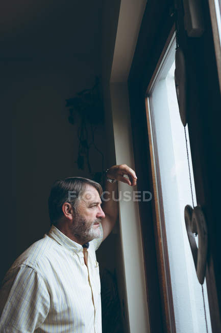 Задумчивый пожилой человек смотрит в окно на дом — стоковое фото