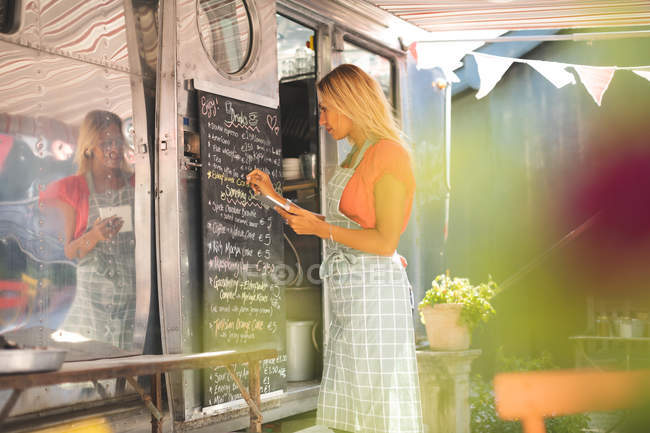 Side view of female waitress writing menu on menu board while using digital tablet — Stock Photo