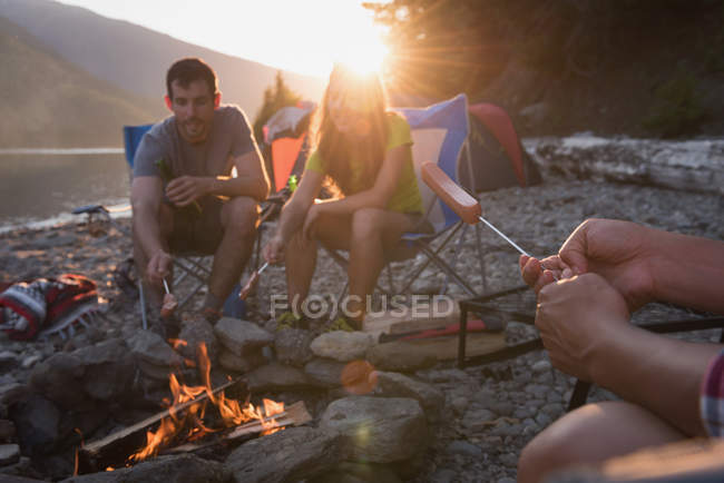 Groupe d'amis chauffage hot dog près de feu de camp — Photo de stock