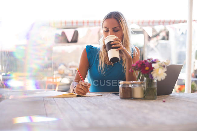 Жінка пише на книзі, маючи каву у відкритому кафе — стокове фото
